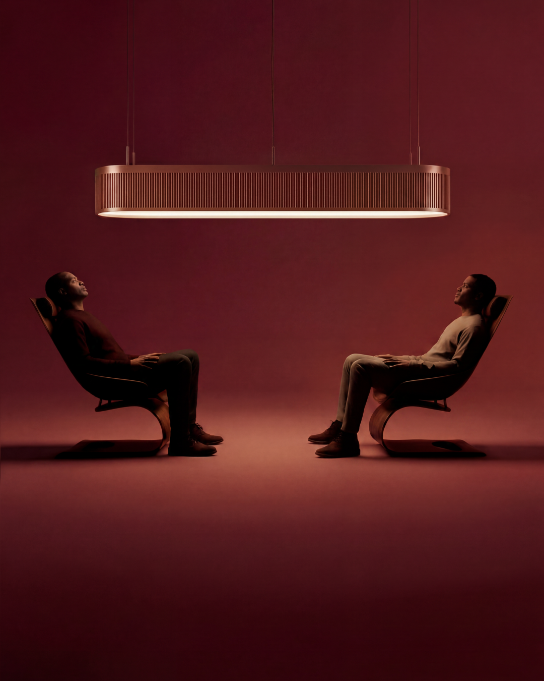 Two figures reclining in curved wooden chairs beneath a long sculptural pendant light, in warm red light